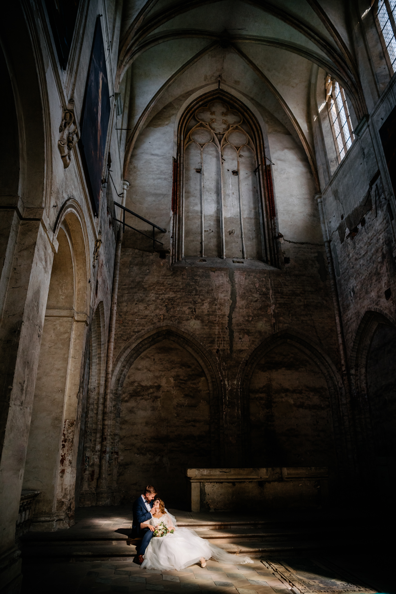 Kobieta i mężczyzna w ślubnych strojach siedzą na schodach w ciemnym gotyckim wnętrzu.